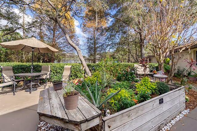 Outdoor garden area with a raised wooden planter filled with various green plants and flowers. There is a patio with a table, chairs, and an umbrella to the left, surrounded by trees and shrubs under a partly cloudy sky.