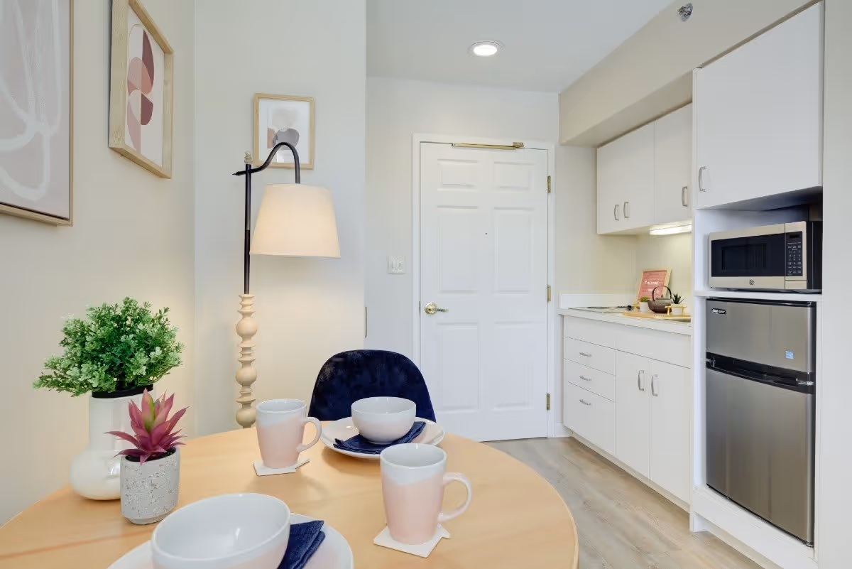 A small dining area with a round wooden table set with white bowls, pink mugs, and navy blue napkins. There are two small potted plants on the table. Behind the table is a tall floor lamp with a beige shade and two abstract framed artworks on the wall. To the right is a compact kitchen area with white cabinets, a microwave, and a small refrigerator. A white door is visible in the background.