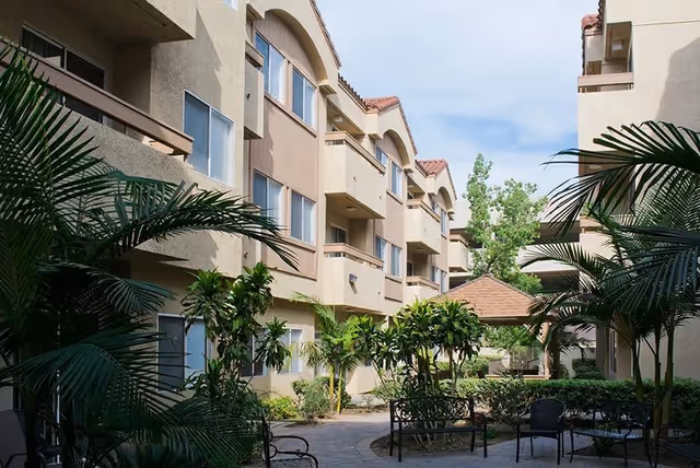 Outdoor courtyard area of a senior living facility with beige multi-story buildings on both sides, lush green plants, palm trees, benches, and a small gazebo under a partly cloudy sky.