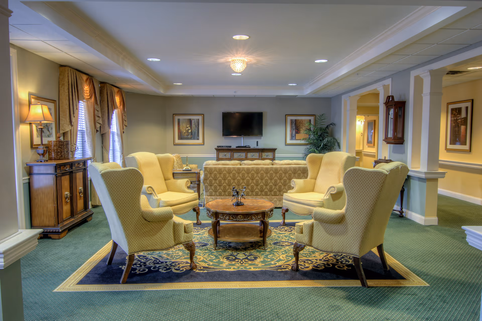 A cozy living room area with four yellow upholstered armchairs arranged around a round wooden coffee table on a decorative rug. Behind the chairs is a patterned sofa facing a wall-mounted flat screen TV. The room has green carpet, framed artwork on the walls, a wooden sideboard with a lamp, and a grandfather clock near an open hallway.