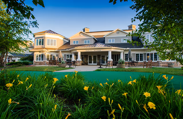Exterior view of Vista Grande Villa, a large two-story building with a covered main entrance, outdoor seating areas, and well-maintained landscaping featuring green grass and yellow flowers under a clear blue sky.