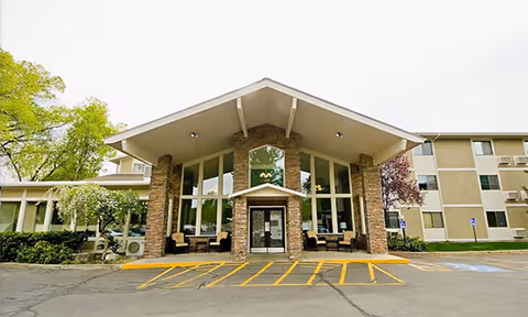 Front exterior view of Williamsburg Retirement Community building with a large peaked roof entrance supported by stone pillars, glass windows, and a paved parking area with yellow lines and handicap parking spaces.