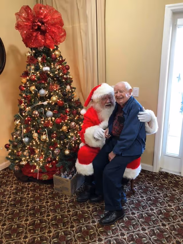 An elderly man sitting on Santa Claus's lap next to a decorated Christmas tree with red and gold ornaments and a large red bow on top, inside a room with patterned carpet and beige walls.