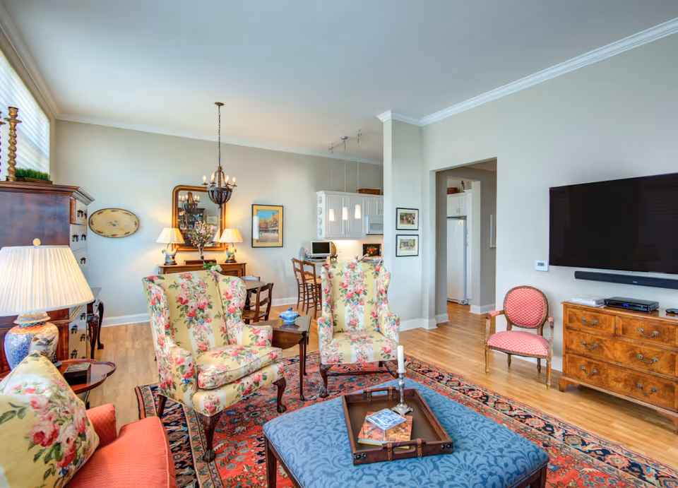 Open living room with floral-upholstered armchairs, a patterned rug and ottoman, a wall-mounted TV, and a view into the dining area and kitchen.