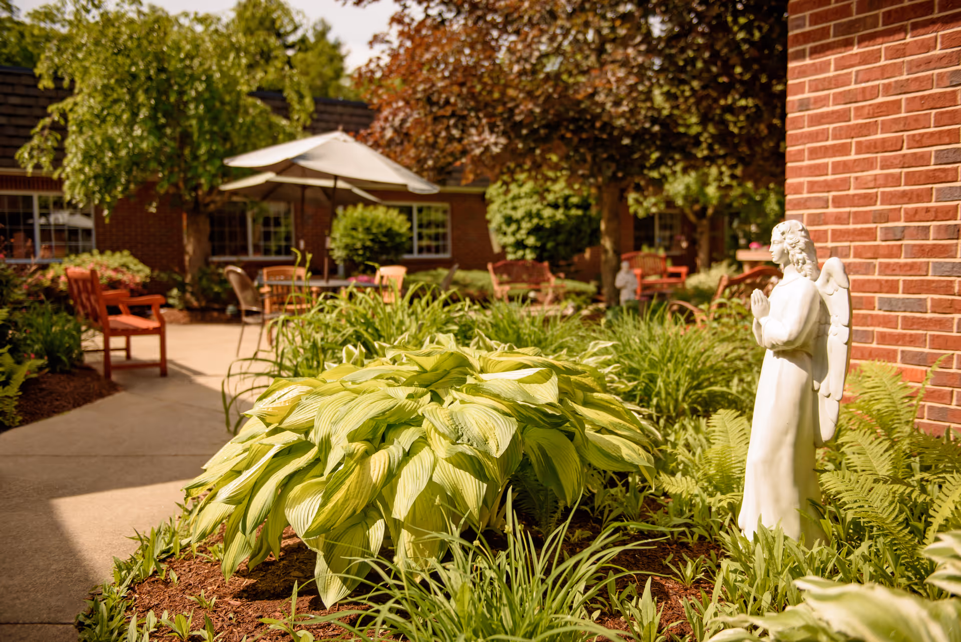 A peaceful outdoor garden area with lush green plants and a white angel statue in the foreground. There are red benches and a table with an umbrella in the background, along with brick buildings and trees providing shade.