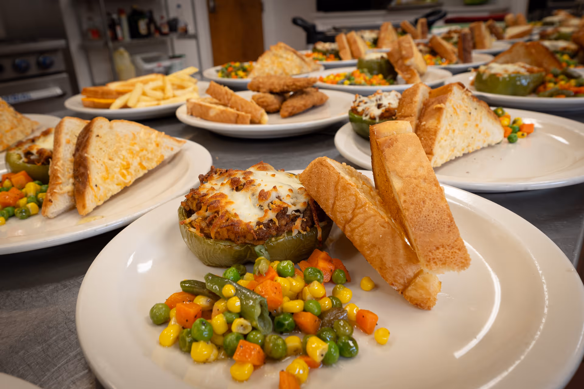 Plates of food arranged on a kitchen counter, each plate featuring a stuffed green bell pepper topped with melted cheese, two slices of toasted bread, and a serving of mixed vegetables including corn, peas, carrots, and green beans.