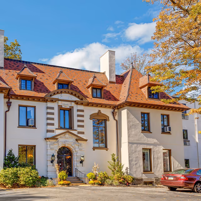 Front exterior of an ornate mansion-style senior living building with a red tiled roof, arched entrance, and landscaped entryway.