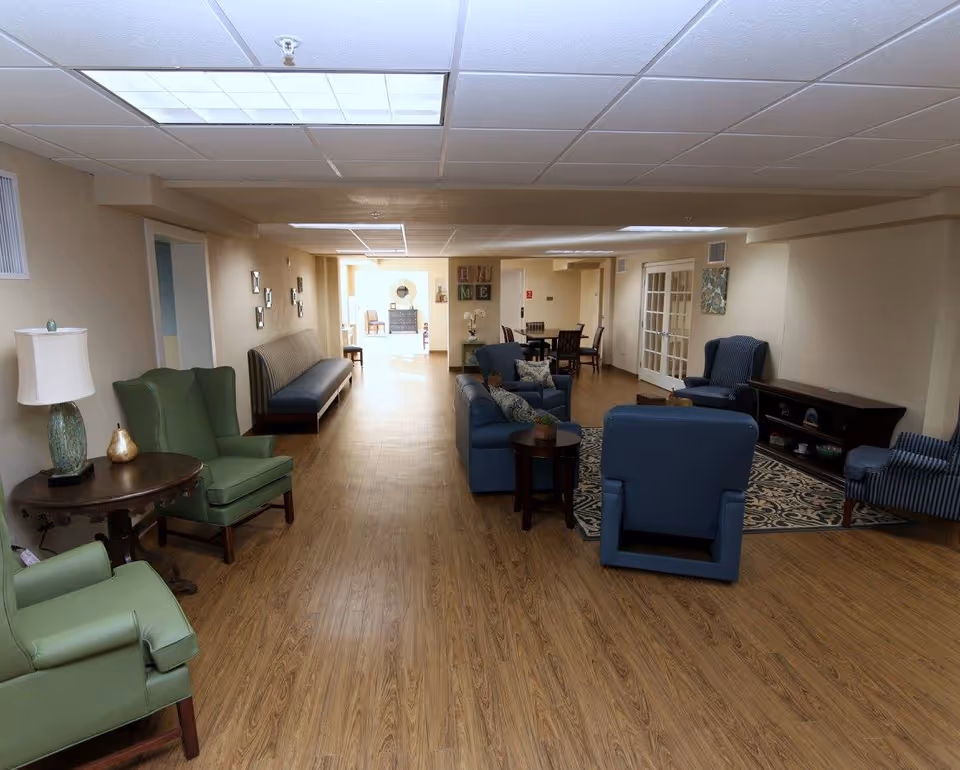 A long communal lounge with armchairs, sofas, tables and wood-look flooring in a senior living facility.