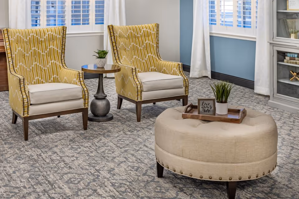 A cozy sitting area with two yellow patterned armchairs separated by a small round wooden table with a potted plant. In front of the chairs is a beige tufted ottoman with a wooden tray holding a small plant and a decorative sign that reads 'Love grows here'. The room has light blue walls, white curtains, and windows with white shutters.