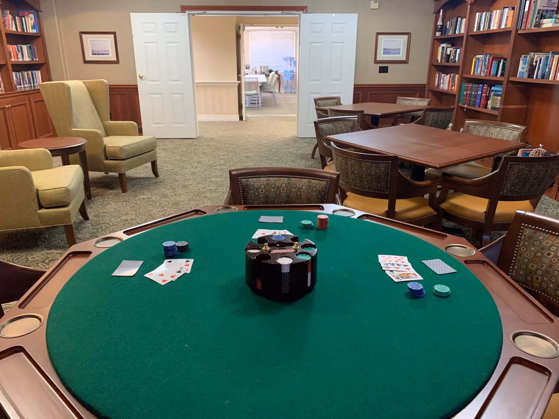 A cozy game room with a green felt poker table in the foreground, featuring playing cards and poker chips. The room has wooden bookshelves filled with books, comfortable armchairs, and tables with chairs. Double white doors lead to another room in the background.