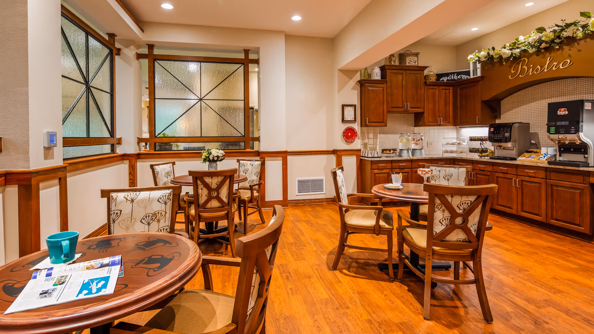 A cozy dining area with wooden floors and several round tables surrounded by cushioned chairs. The room features a small kitchenette with wooden cabinets, a coffee machine, and a water dispenser. Decorative elements include a floral arrangement on one table and a sign above the kitchenette that reads 'Bistro'.