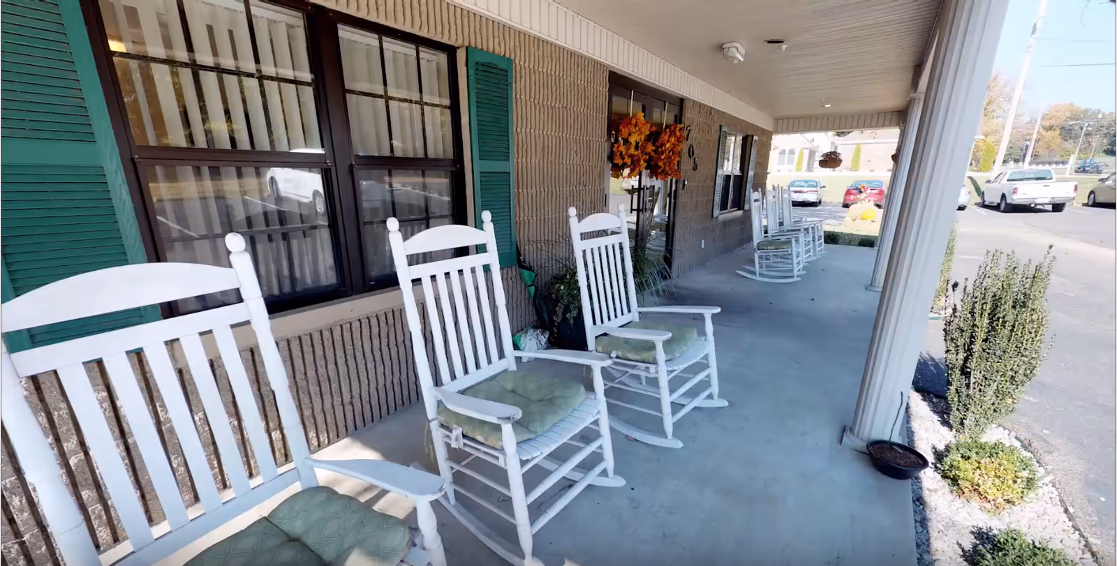 A covered outdoor porch area with several white wooden rocking chairs with green cushions lined up along the wall of a building. The building has green shutters and windows with curtains. There are some plants and a fall-themed wreath near the entrance door. In the background, a parking lot with several cars is visible.