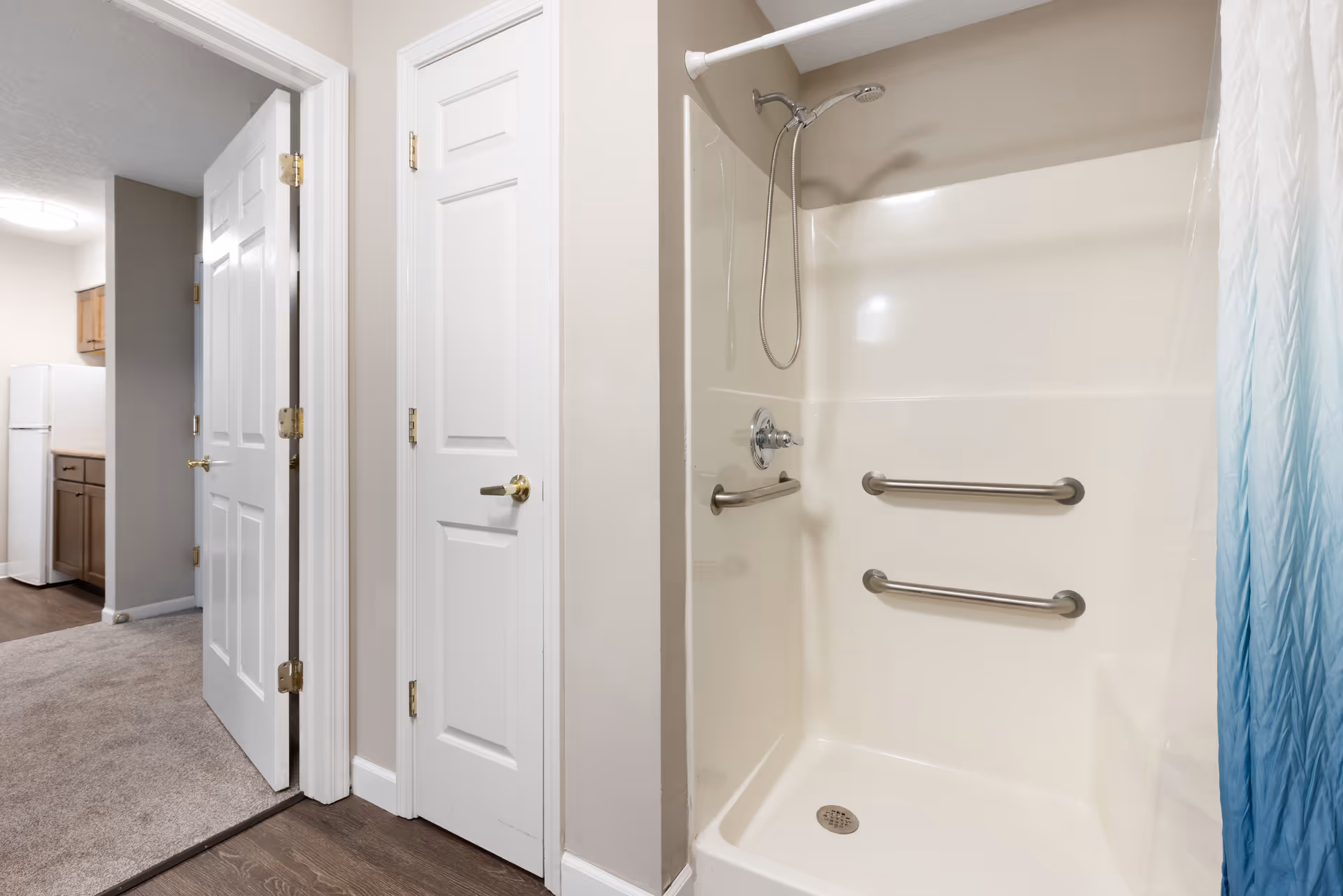 Interior view of a senior living facility bathroom shower area with two horizontal grab bars and a handheld showerhead. Adjacent to the shower are two closed white doors, and a hallway leading to a kitchen area with a refrigerator and wooden cabinets.