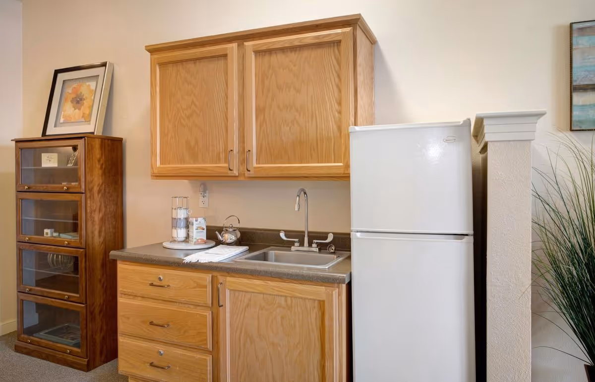 Small kitchenette with oak cabinets, a sink and countertop, a white mini refrigerator, and a glass-front shelving unit.