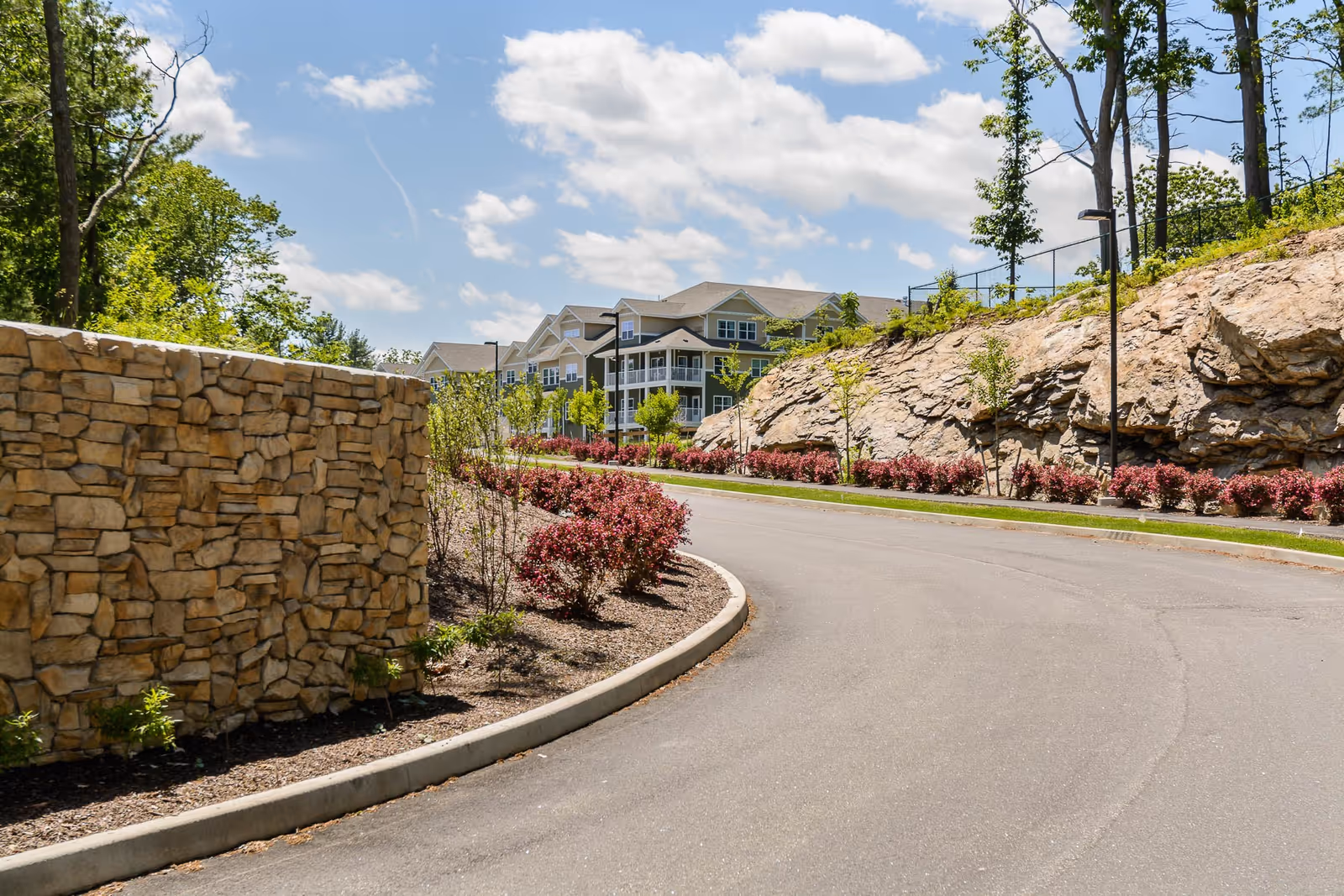 Curved paved driveway lined with landscaped bushes and small trees, with a stone wall on the left and rocky hillside on the right. In the background, a multi-story residential building with balconies is visible under a partly cloudy blue sky.