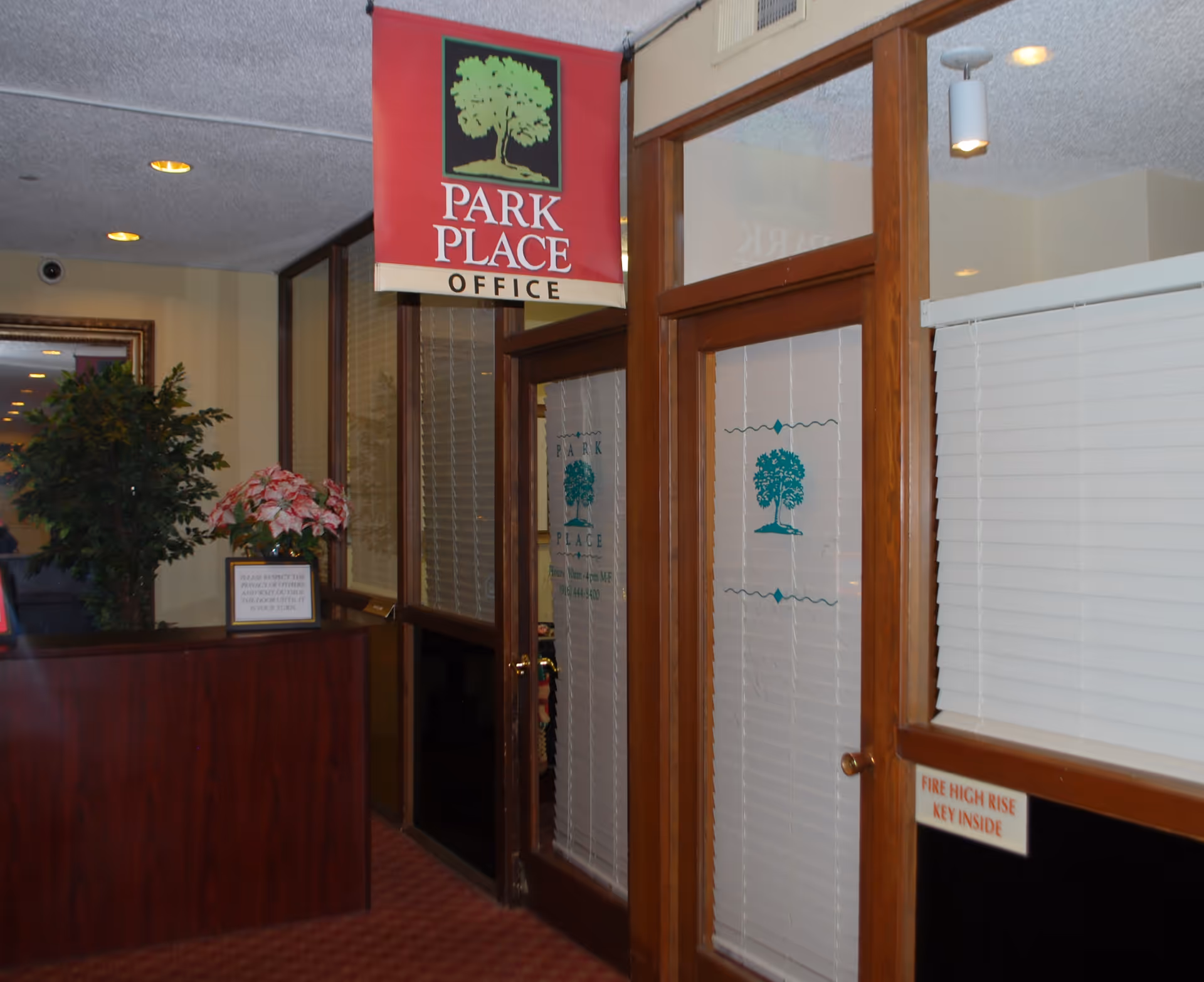Interior view of a reception area showing a 'Park Place Office' hanging sign, a wood-framed glass office door with blinds, and potted plants.