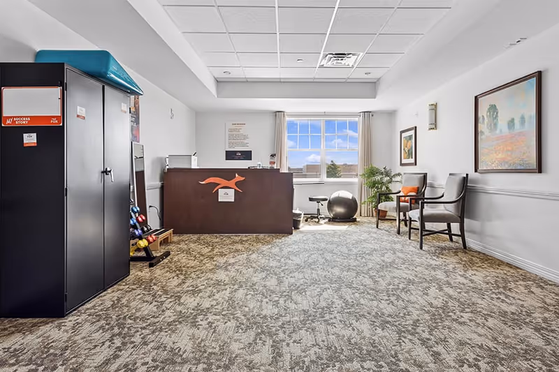 A bright room with a large window showing a blue sky with clouds. The room has a patterned carpet and white walls decorated with framed paintings. On the left side, there is a black cabinet with a blue top and a rack holding colorful dumbbells. In the center, there is a dark wooden reception desk with an orange kangaroo logo. On the right side, there are two chairs with cushions and a potted plant between them. The ceiling has white tiles with fluorescent lighting.