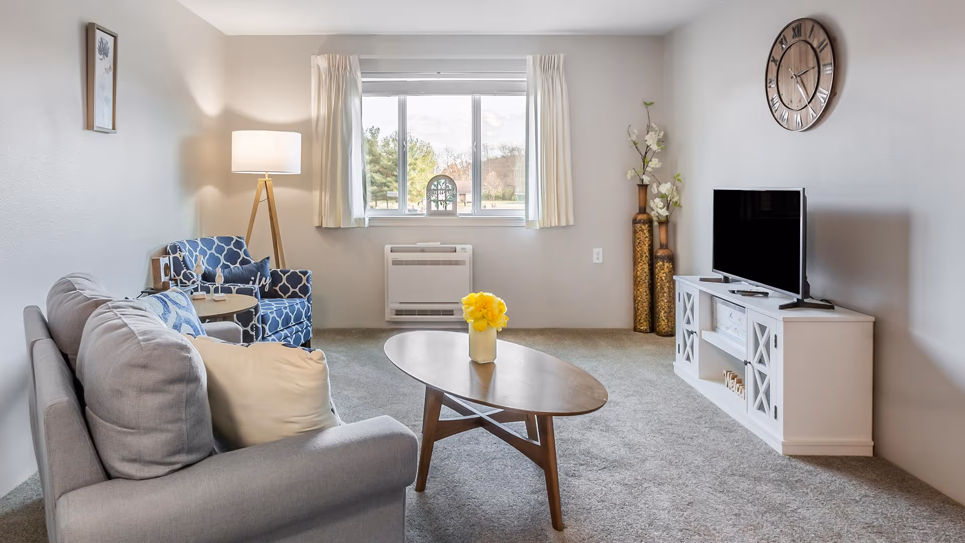 A cozy living room with a gray sofa and a beige pillow, a blue patterned armchair, a wooden coffee table with a vase of yellow flowers, a floor lamp, a window with white curtains, a wall clock, a flat-screen TV on a white TV stand, and decorative tall vases with flowers in the corner.