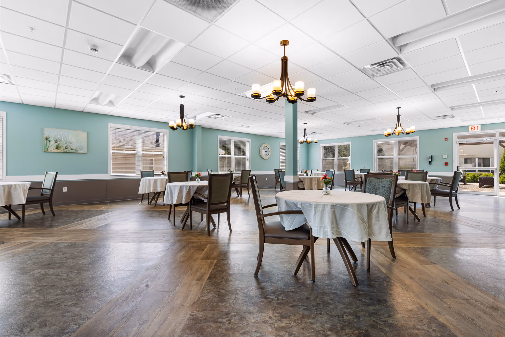 A spacious dining room with round tables covered in white tablecloths and wooden chairs arranged around them. The walls are painted light blue with large windows letting in natural light. Several chandeliers hang from the white tiled ceiling. There is a clock on one wall and an exit door leading outside.