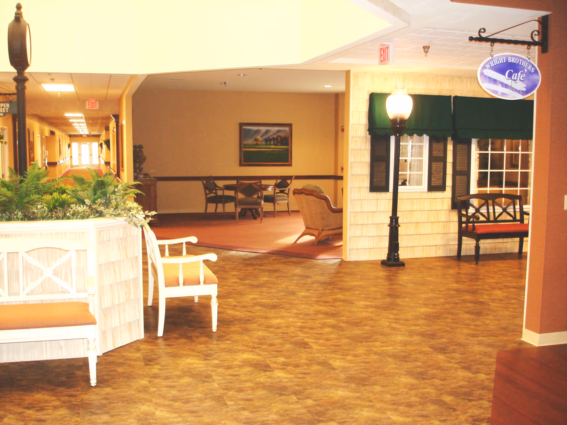 Interior common area of a senior living facility with seating, plants, a hallway, and a hanging 'Wright Brothers Cafe' sign.