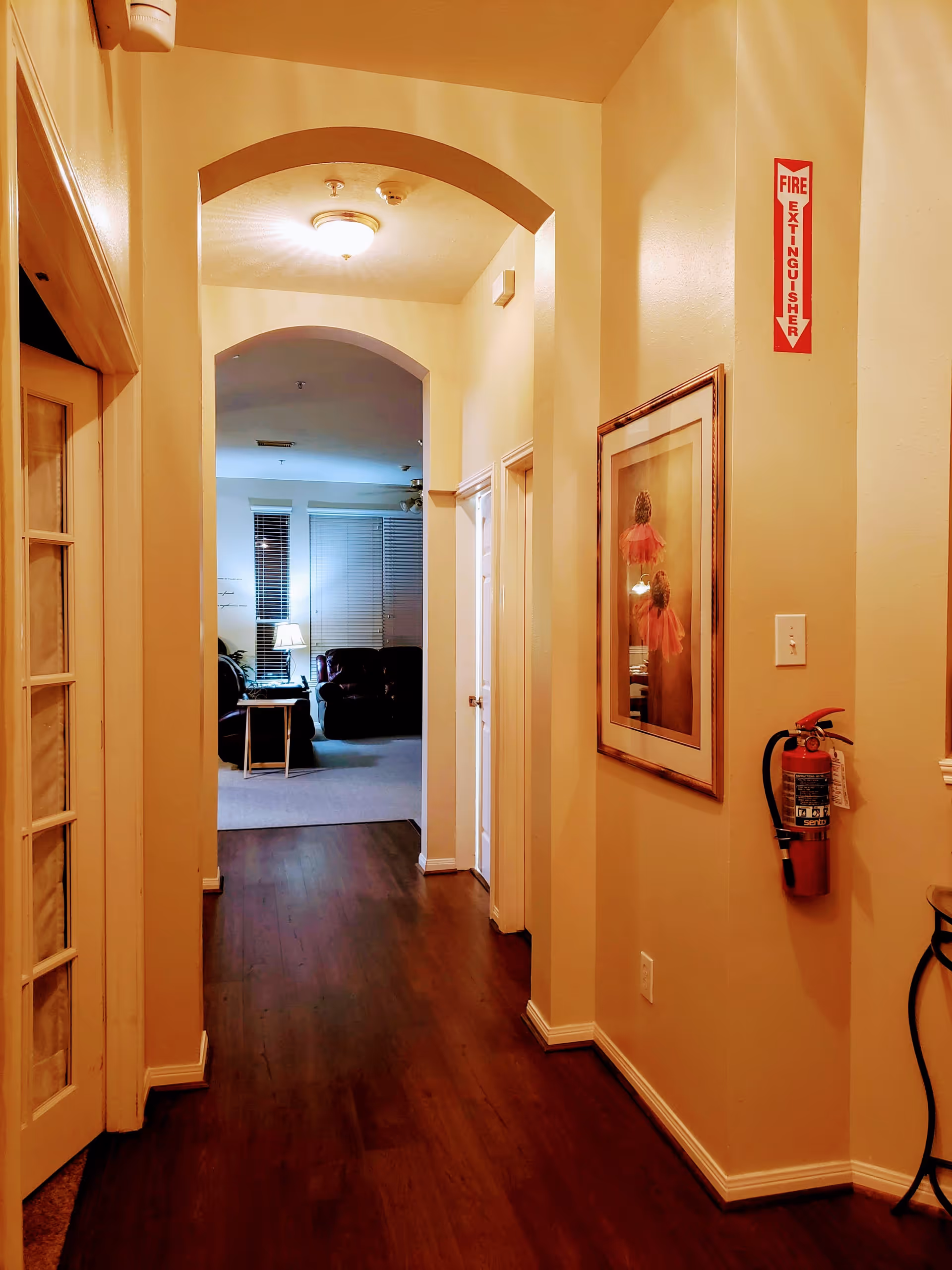A hallway with wooden flooring and cream-colored walls leading to a living room area with a lamp and leather chairs. The hallway has two arched doorways, a framed picture on the right wall, and a fire extinguisher mounted below a fire extinguisher sign.
