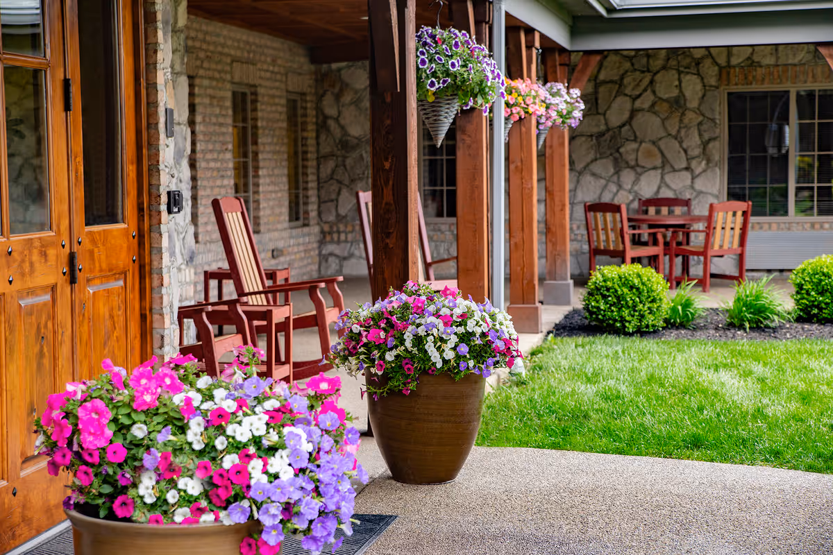 Covered outdoor patio area with wooden rocking chairs and a table with chairs. There are hanging flower baskets and large flower pots with colorful flowers. The building exterior is made of stone and brick with wooden doors and windows.