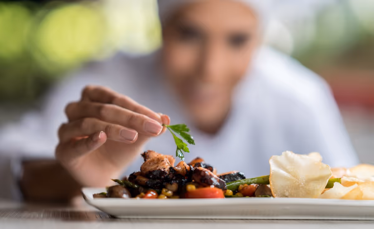 A chef garnishing a plated dish with a fresh green herb, with the focus on the food and the chef's hand, while the chef's face is blurred in the background.