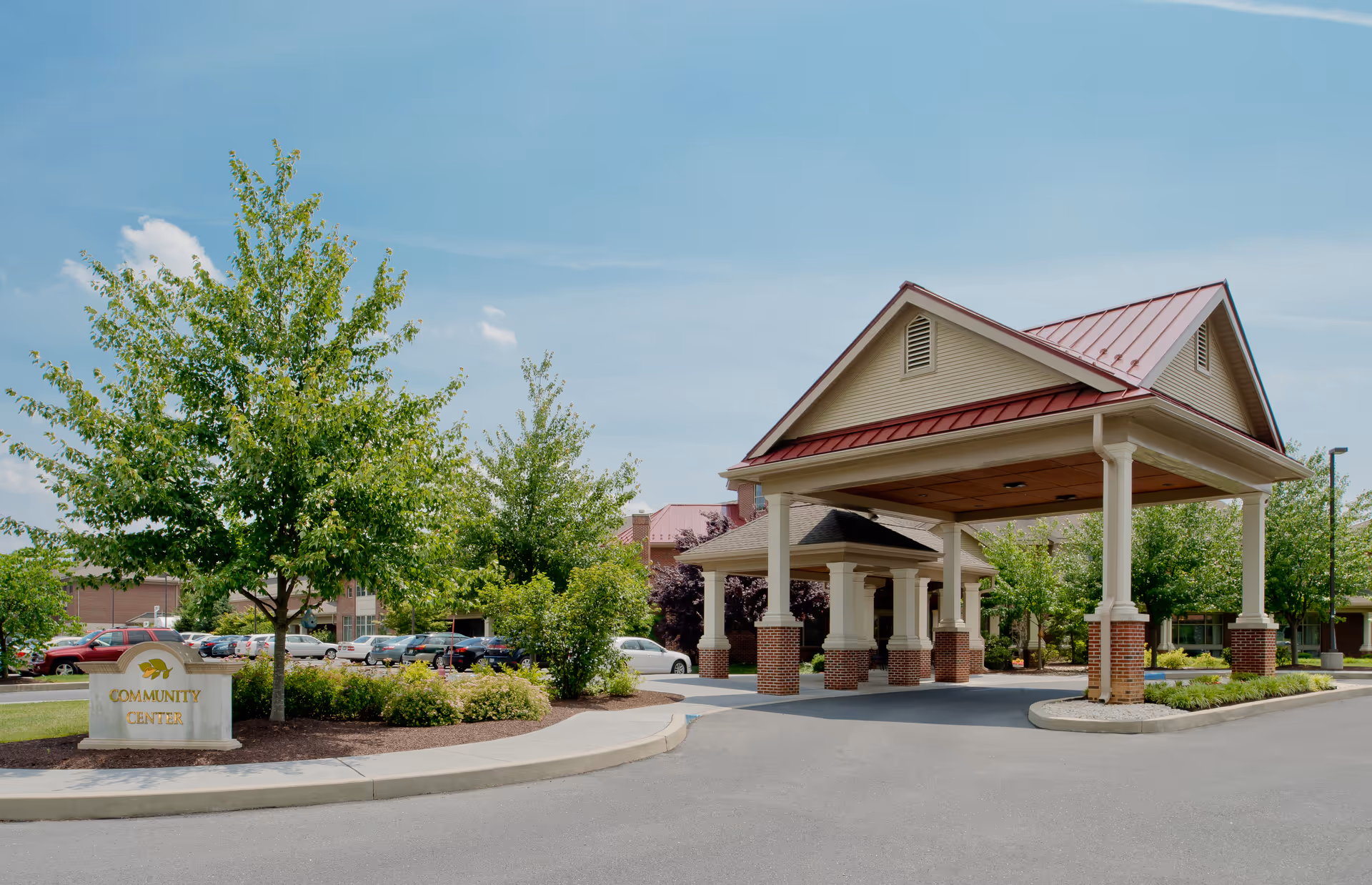 Entrance to a community center with a covered drop-off area supported by white columns with brick bases, surrounded by trees and landscaping. A sign near the curb reads 'Community Center'. Several parked cars are visible in the background under a clear blue sky.