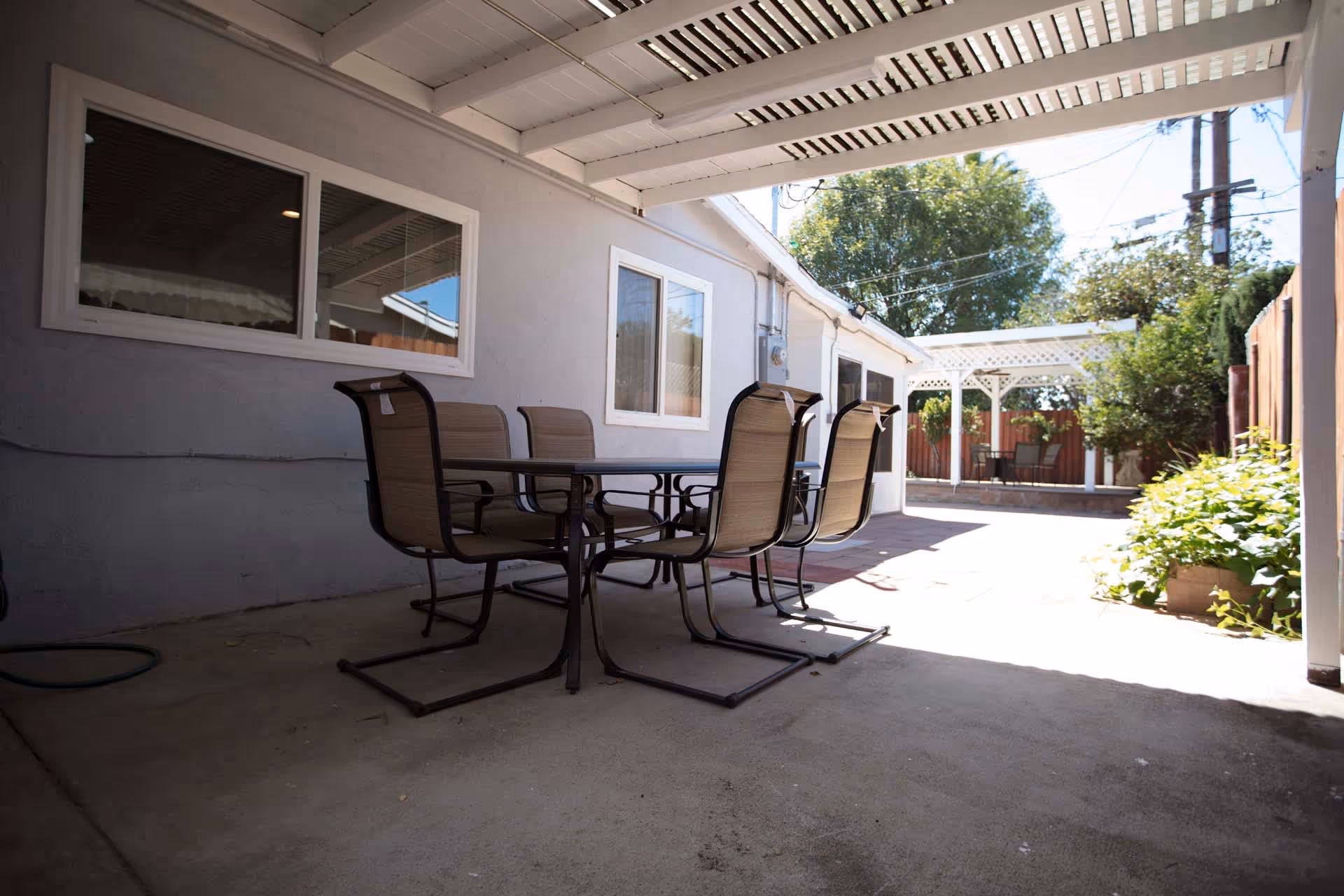 Outdoor patio area with a covered section featuring a table and four chairs. The patio extends to an uncovered area with a white pergola and additional seating. Surrounding the patio are plants, trees, and a wooden fence.
