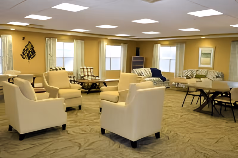 A spacious living room area with beige walls and carpeted floor. The room contains several cream-colored armchairs arranged in a circle, a few patterned sofas, and a wooden table with chairs. Large windows with light curtains allow natural light to enter the room. The ceiling has recessed lighting panels.