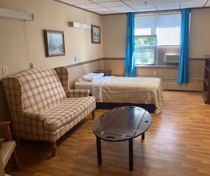 Small furnished senior living bedroom with a single bed, plaid loveseat, coffee table, and a window with blue curtains and an air conditioner.