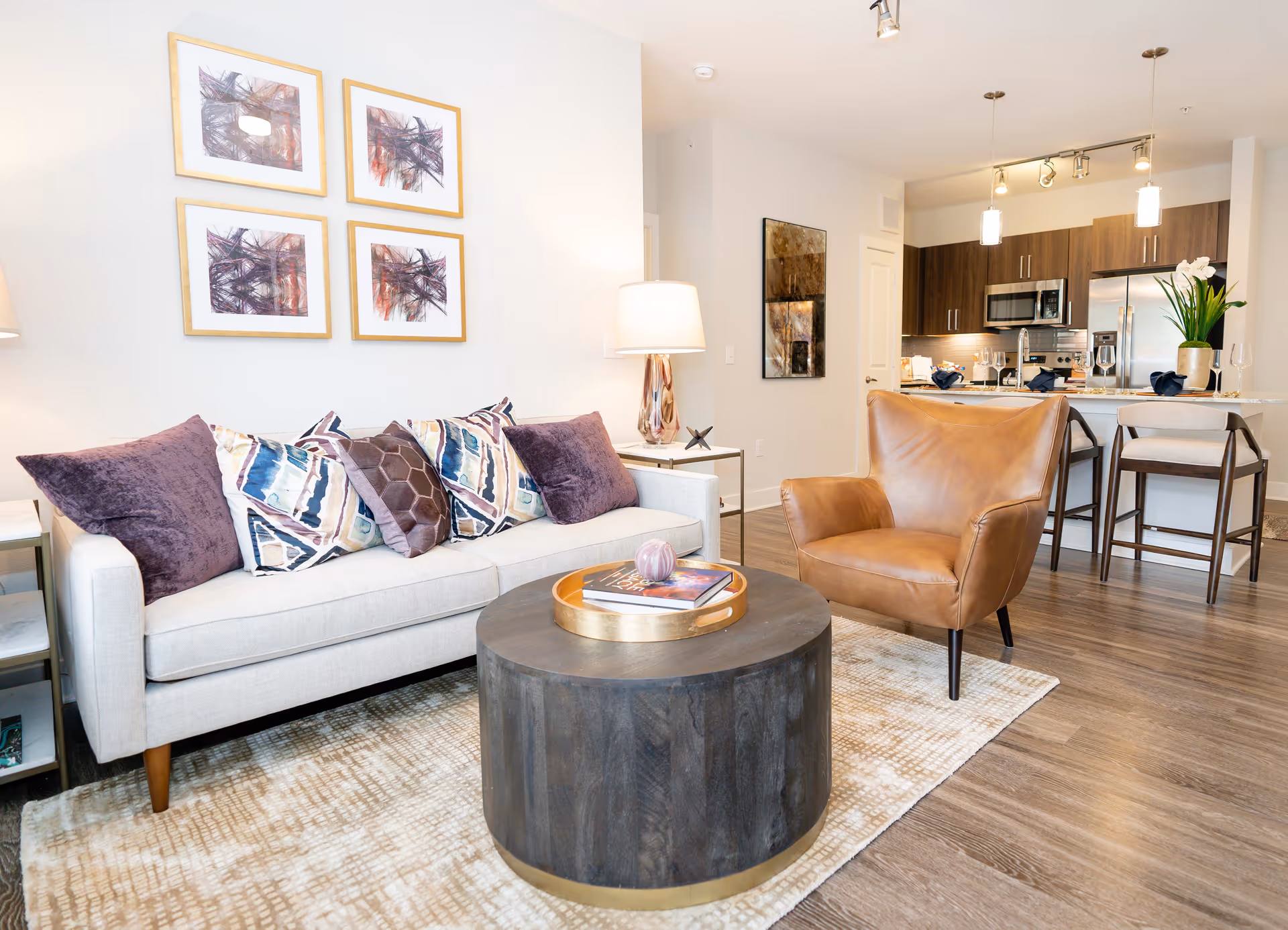 A modern living room with a light beige sofa adorned with patterned and solid purple pillows, a round dark wooden coffee table with a decorative tray and book, a brown leather armchair, and a light-colored area rug. In the background, there is a kitchen area with dark wood cabinets, stainless steel appliances, a kitchen island with bar stools, and pendant lighting. The walls are decorated with framed abstract artwork and a lamp on a side table next to the sofa.