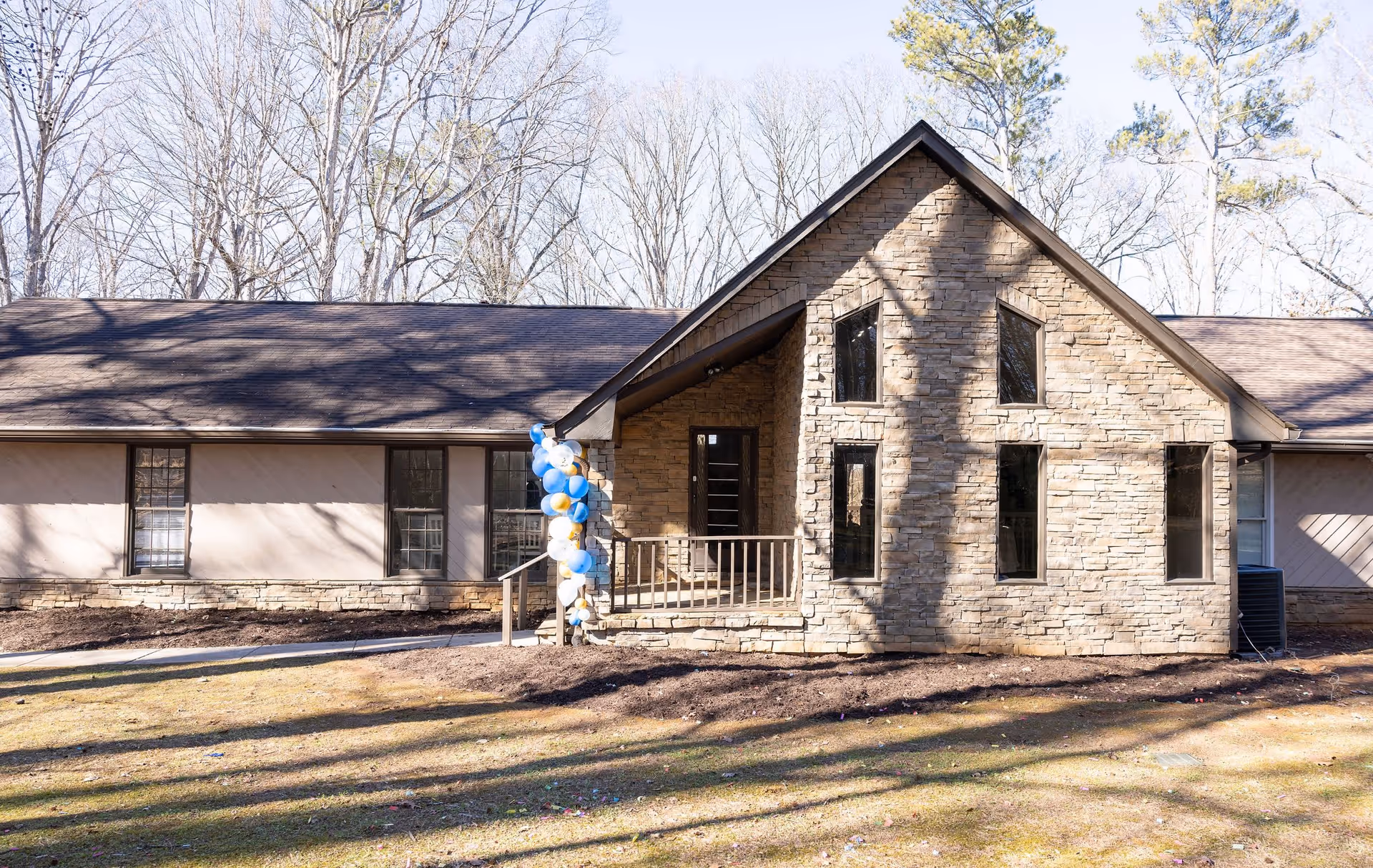 Exterior view of a single-story senior living facility building with stone and beige siding, several windows, a small porch decorated with blue, white, and gold balloons, and leafless trees in the background.