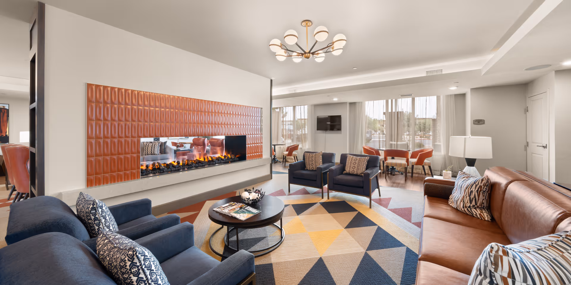 A modern living room with a large brown leather sofa, two blue armchairs, and a blue loveseat arranged around a round coffee table on a colorful geometric rug. A contemporary fireplace with orange tile surround is built into the wall. Large windows with sheer curtains allow natural light to fill the space. A flat-screen TV is mounted on the far wall, and a chandelier with multiple round lights hangs from the ceiling.