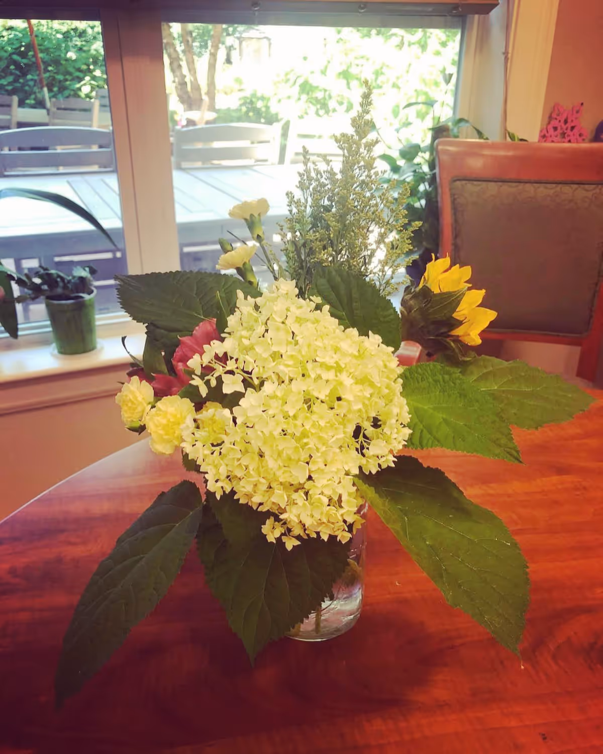 A bouquet of mixed flowers including white hydrangeas, yellow sunflowers, and other small flowers arranged in a glass jar on a wooden table. In the background, there is a window with a view of an outdoor patio with chairs and greenery.