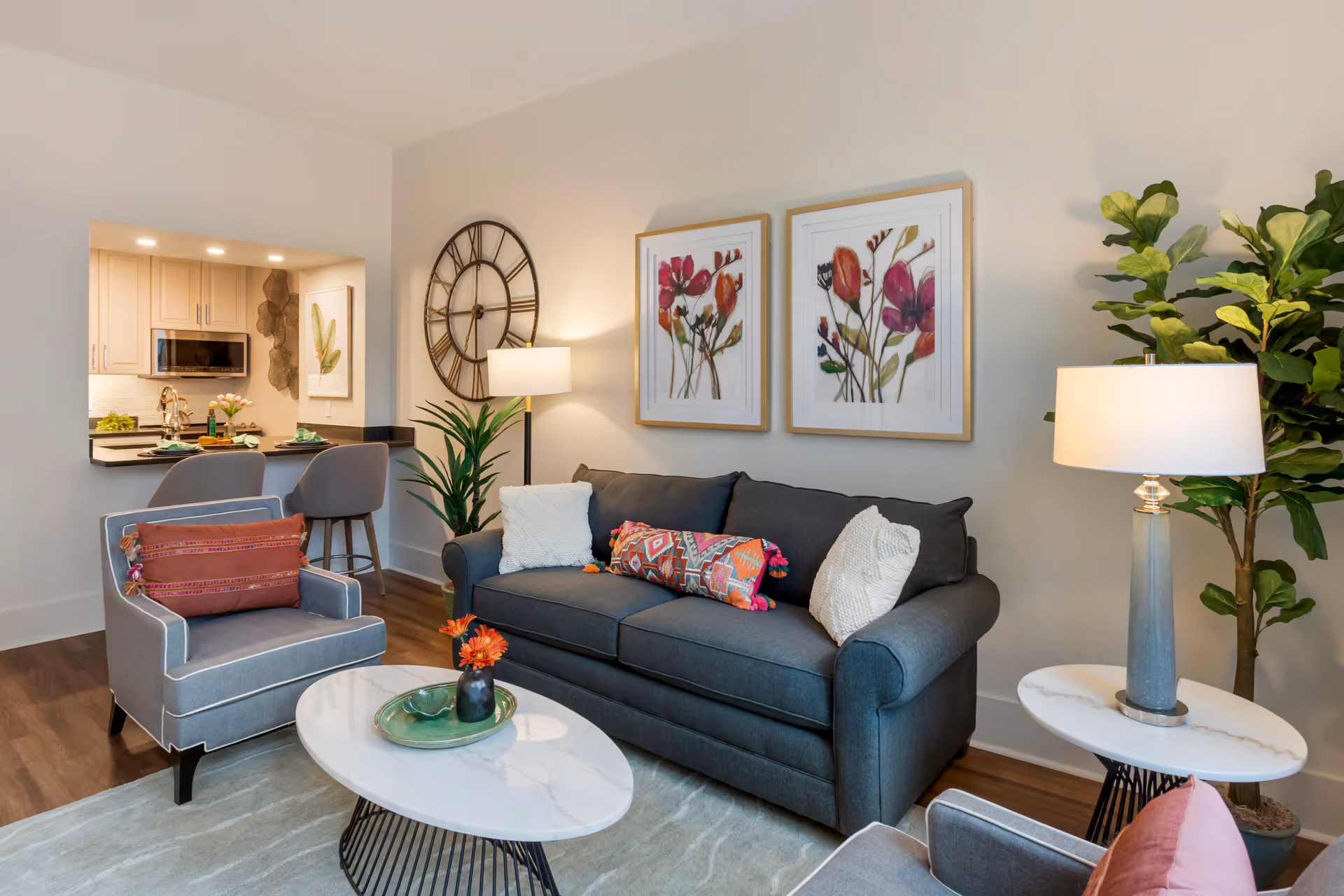 A cozy living room with a dark gray sofa adorned with colorful and white pillows, two gray armchairs with decorative pillows, a round white marble coffee table with a green tray and orange flowers, a side table with a lamp, two framed floral artworks on the wall, a large decorative wall clock, a tall green plant, and a view into a kitchen area with bar stools.