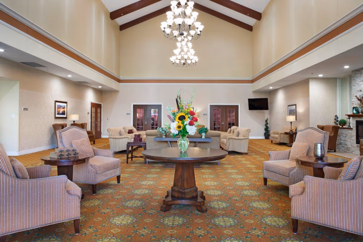 Spacious senior living lounge with upholstered chairs arranged around a central table holding a floral arrangement, under a vaulted ceiling with chandeliers.