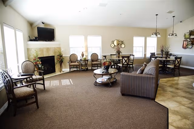 A spacious living room area with beige walls and carpeted floor. The room features a fireplace with a TV mounted above it, several chairs and tables arranged around the room, a striped sofa, and decorative plants and wall art. Large windows allow natural light to fill the space.