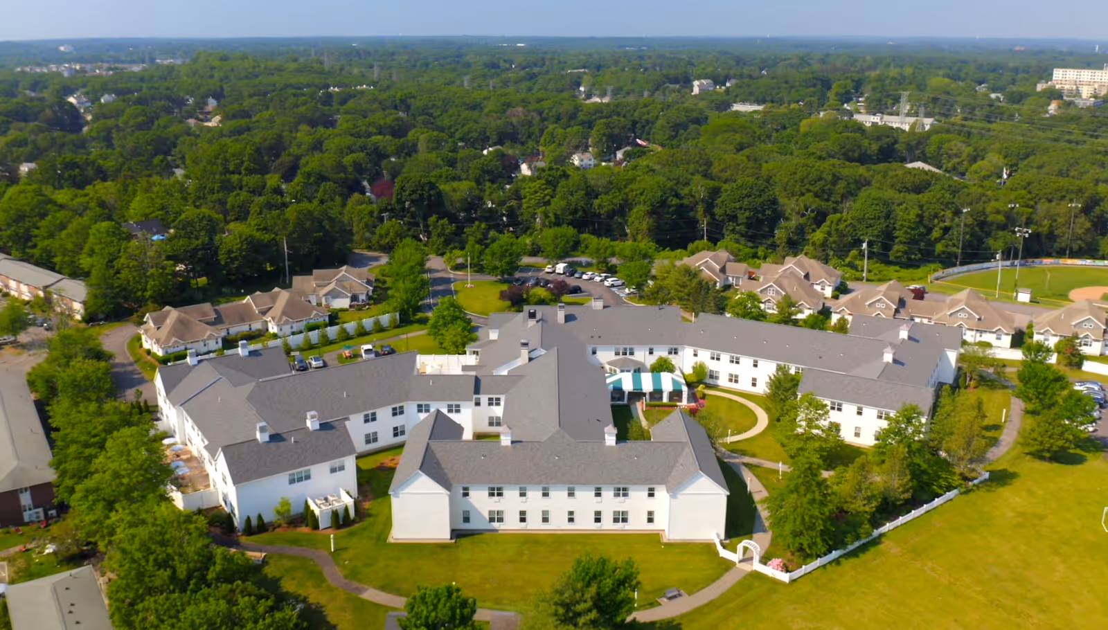 Aerial view of a large senior living facility named Allerton House Weymouth surrounded by green trees and lawns, with nearby residential houses and a baseball field visible in the background.