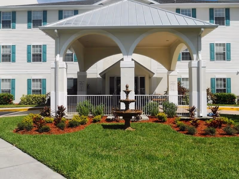 Outdoor view of a white multi-story building with green window shutters behind a covered pavilion structure. In front of the pavilion is a tiered water fountain surrounded by landscaped bushes and green grass.