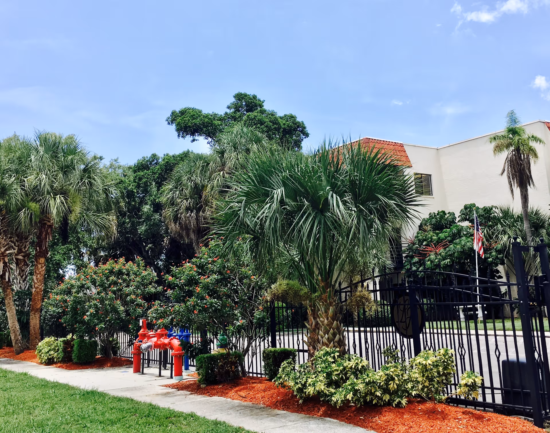 Exterior view of a building with a white facade and red roof tiles, surrounded by lush greenery including palm trees and bushes. A black metal fence with a gate is in front, along with red and blue fire hydrants. The sky is clear and blue.