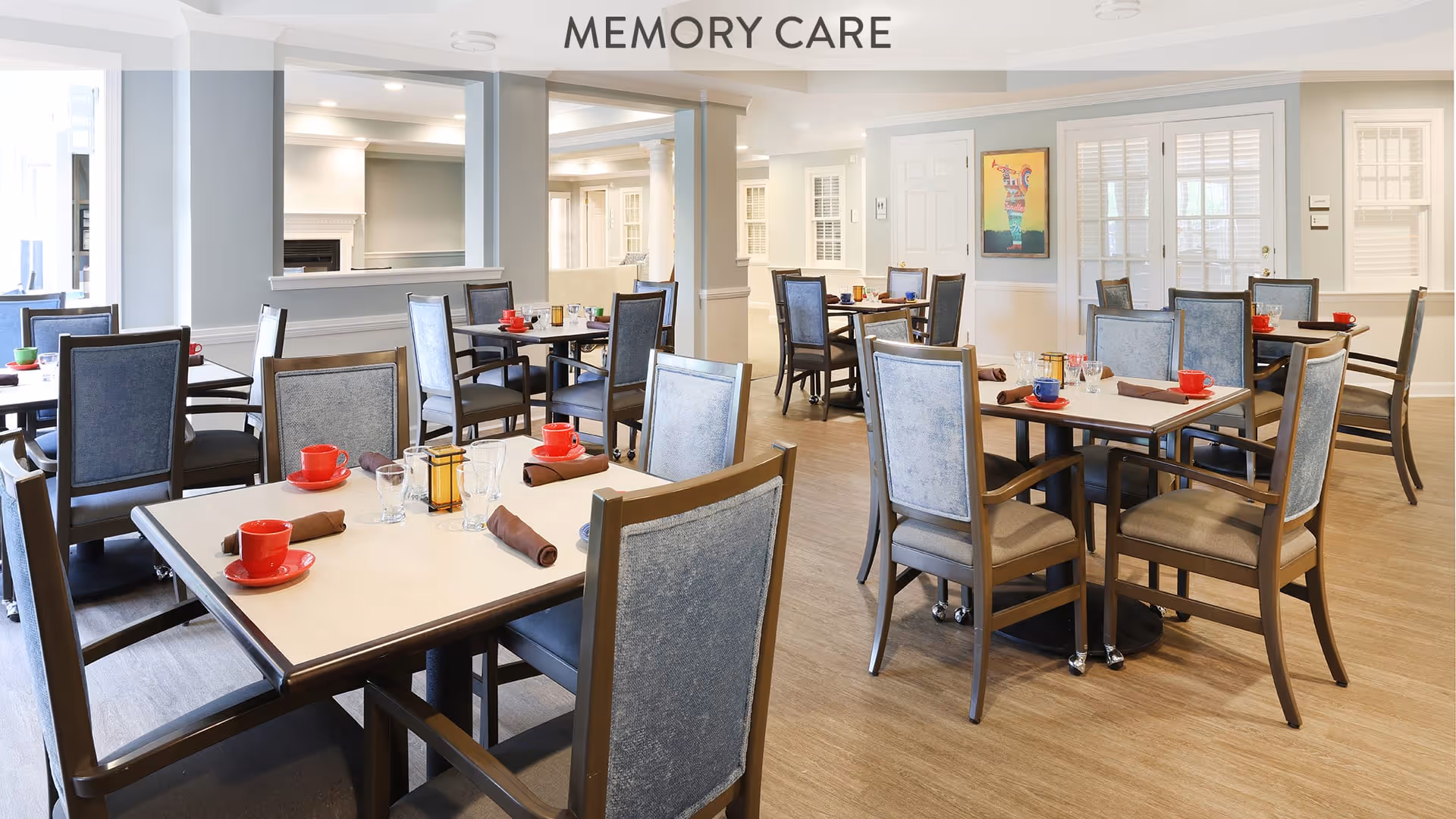 A bright and spacious dining room with multiple tables and chairs arranged neatly. Each table is set with red cups, glasses, and brown napkins. The room has light-colored walls, large windows with white shutters, and a sign above that reads 'MEMORY CARE'.