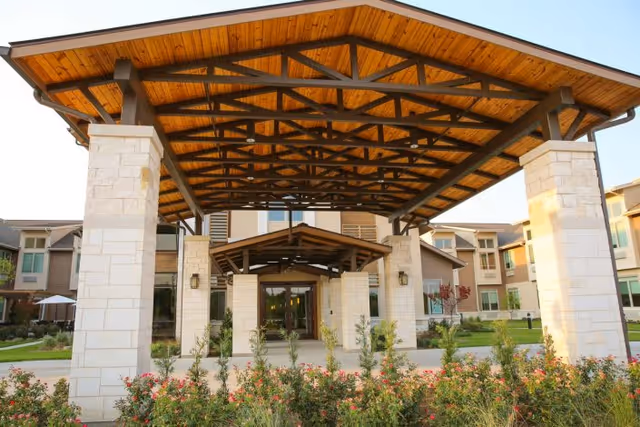 Entrance of a senior living facility with a large wooden canopy supported by stone pillars, surrounded by landscaped greenery and flowers, with multi-story residential buildings in the background.