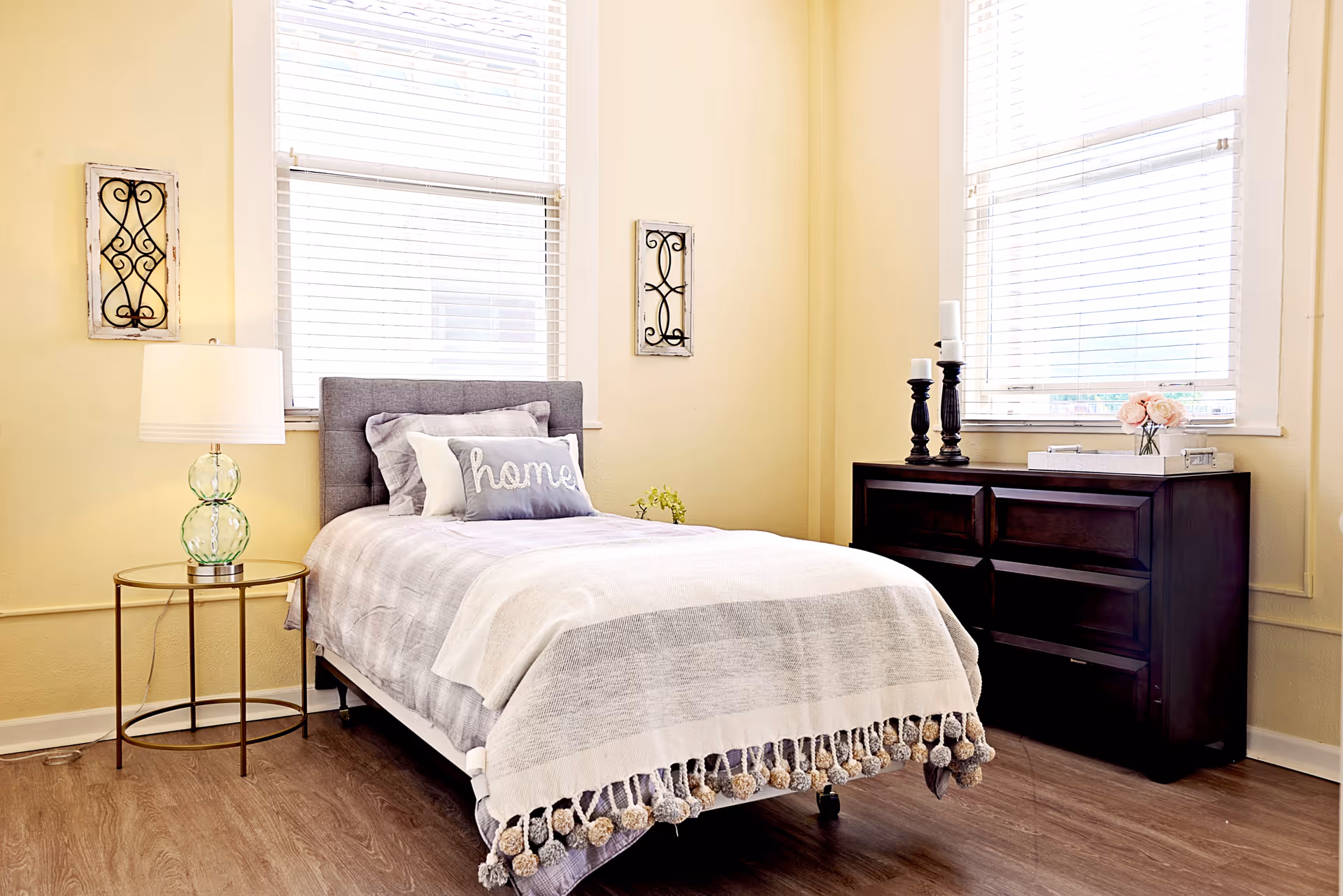 A cozy bedroom with a single bed featuring a gray upholstered headboard, white and gray bedding, and a decorative pillow with the word 'home'. There is a round side table with a glass lamp on the left side of the bed. On the right side, there is a dark wooden dresser with two black candle holders and a small vase with pink flowers. The room has two large windows with white blinds, light yellow walls, and wooden flooring.