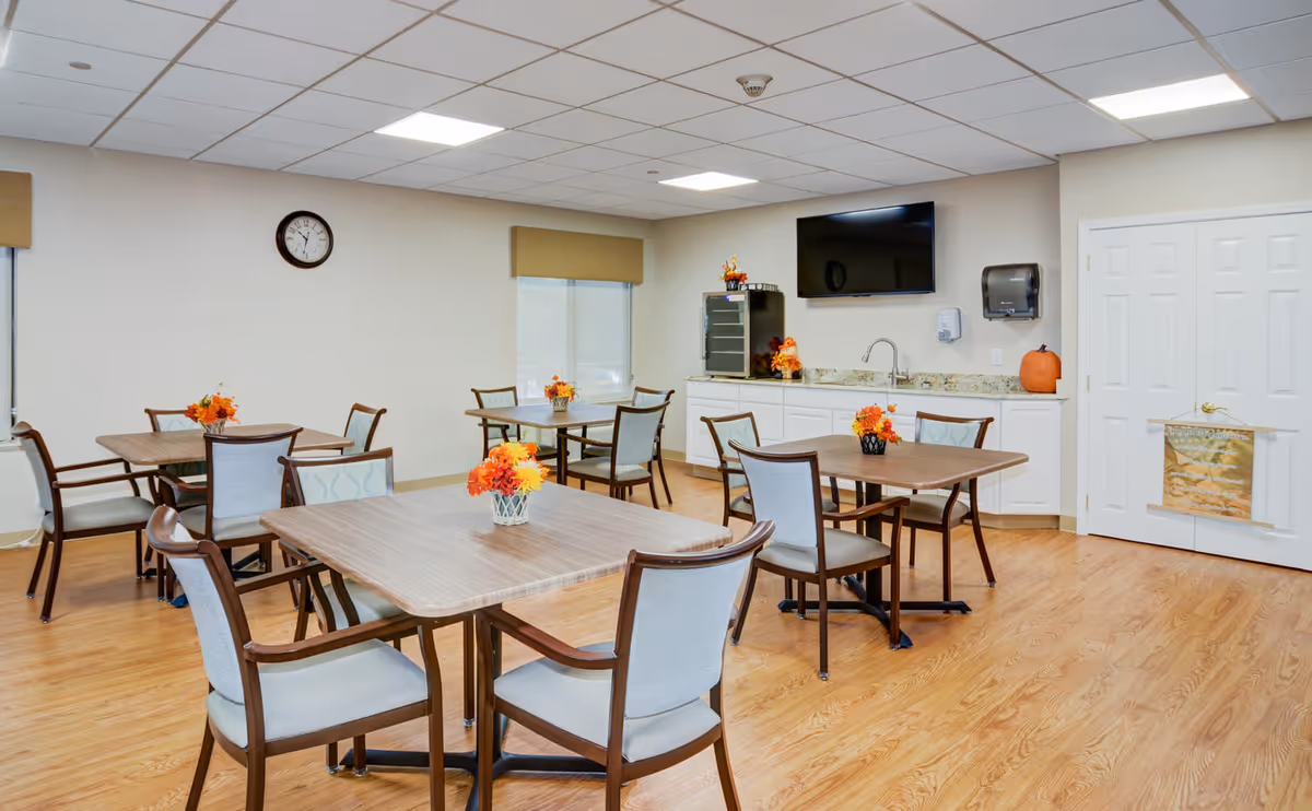 Bright communal dining room with multiple tables and chairs, a small kitchenette with a wall-mounted TV, and floral centerpieces.