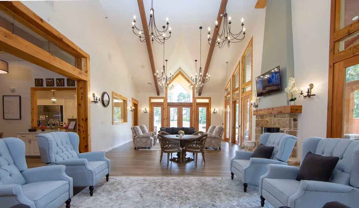 A spacious, well-lit living room with high vaulted ceilings featuring wooden beams and multiple chandeliers. The room has light blue upholstered armchairs with dark cushions, a stone fireplace with a mounted TV above it, and a round wooden table surrounded by chairs in the center. Large windows and glass doors allow natural light to fill the space, and there is a view into an adjacent kitchen area with white cabinetry.