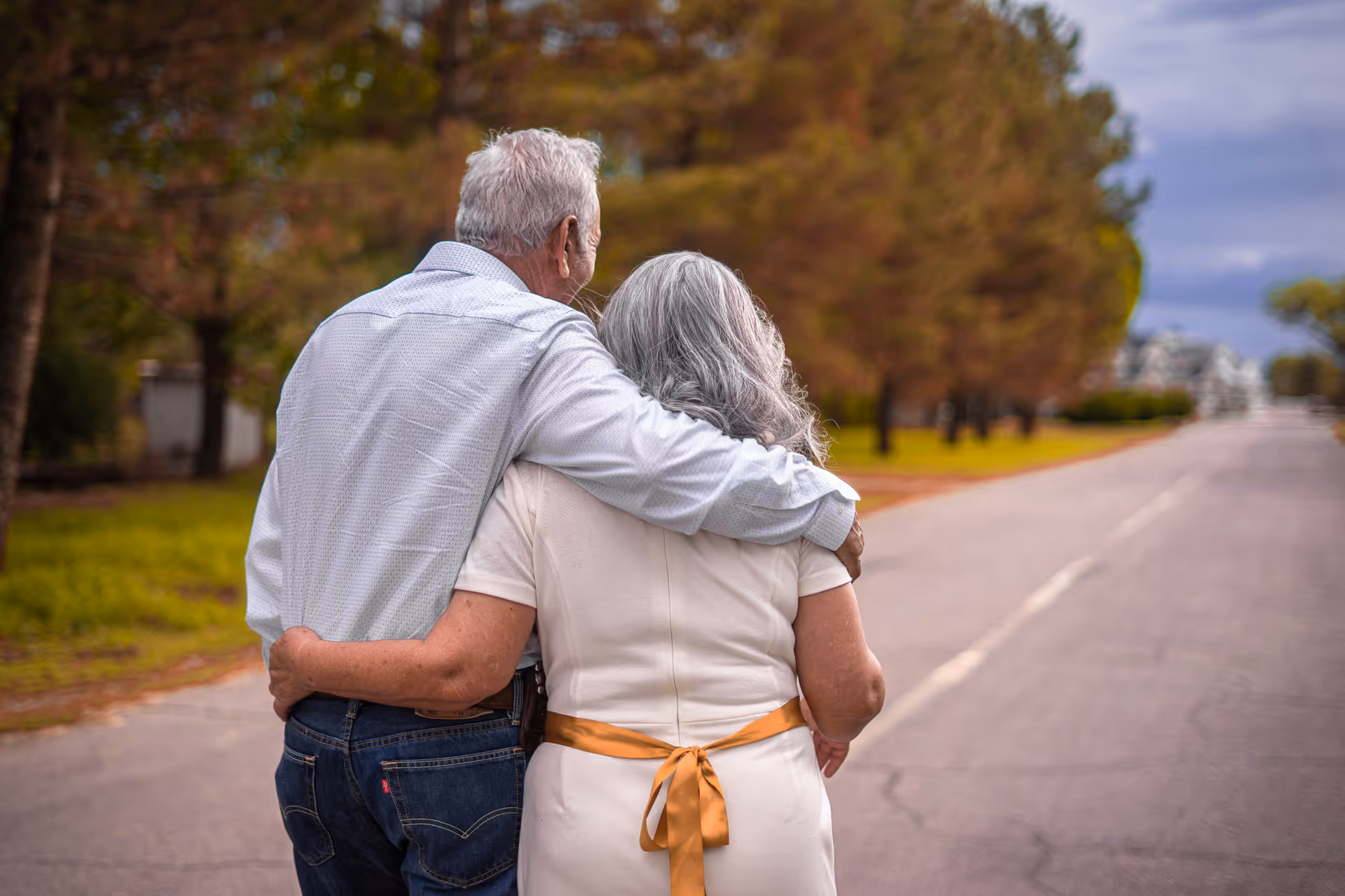 An elderly couple walking arm in arm down a tree-lined road, viewed from behind. The man is wearing a light blue shirt and jeans, and the woman is wearing a white dress with a yellow ribbon tied at the back. The scene is outdoors with greenery and trees on the side of the road.