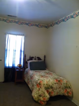 A small bedroom with a single bed covered in a floral bedspread. There is a wooden headboard and a large pillow on the bed. Next to the bed is a wooden nightstand with a small lamp and a decorative item. A window with sheer white curtains and darker drapes is on the left wall. The walls are white with a floral border near the ceiling.