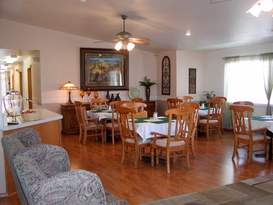 A dining room in an assisted living facility with several wooden tables and chairs arranged neatly. Each table is covered with a white tablecloth and a green runner. The room has wooden flooring, a ceiling fan with lights, and a large window with sheer green curtains letting in natural light. There is a hallway visible to the left and decorative items including a framed painting, a lamp, and vases on a sideboard against the wall.