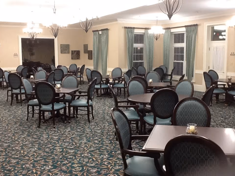 A spacious dining room with multiple round tables surrounded by blue cushioned chairs. The room has patterned carpet flooring, beige walls, green curtains on the windows, and several hanging light fixtures providing warm lighting.